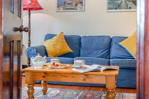 A cozy living room with a blue sofa, a wooden coffee table with tea cups and a teapot, and a door with a red lamp. - 3 Bed in Padstow (oc-p00043) (Saint Merryn)