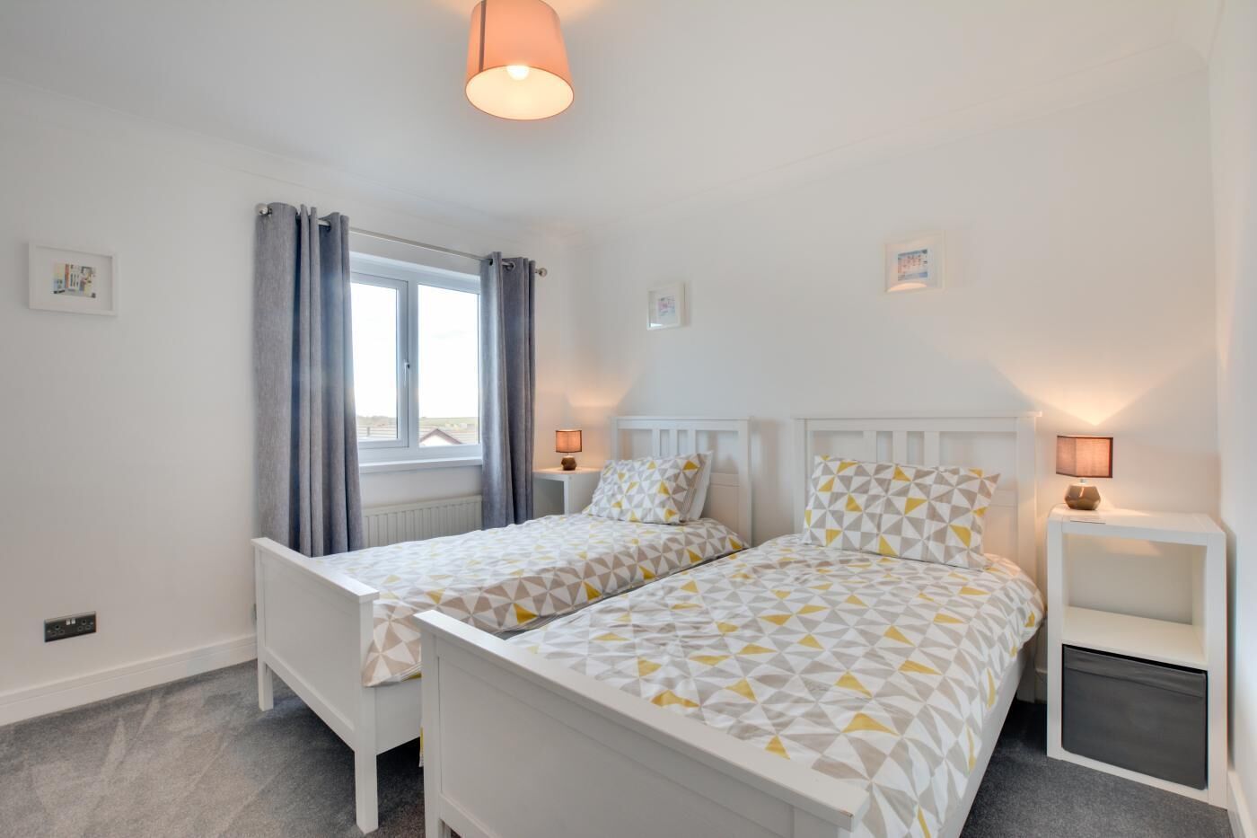 Een slaapkamer met twee enkele bedden, een raam met gordijnen, nachtlampjes en een klein tafeltje.