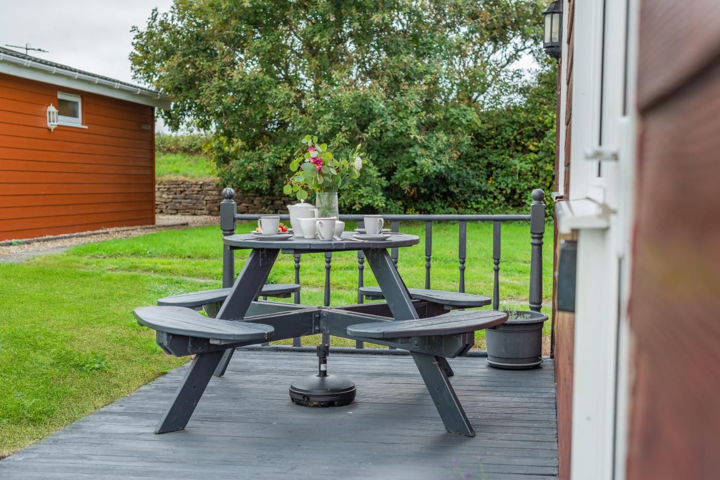 A wooden deck with a round table and benches, a small potted plant, and a white building in the background.