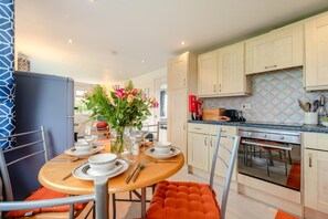 A modern kitchen with a dining area, featuring a table set for four, a vase with flowers, and a red fire extinguisher. - 2 Bed in Padstow (oc-p00155) (Penrose)