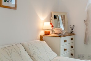 A bedroom with a bed, a bedside table with a lamp, a mirror, and a dresser with drawers. - 2 Bed in Padstow (oc-p00155) (Penrose)