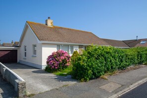 A single-story house with a sloped roof, a garage, and a well-maintained garden with a large flowering bush. - 2 Bed in Padstow (oc-p00464) (Saint Merryn)