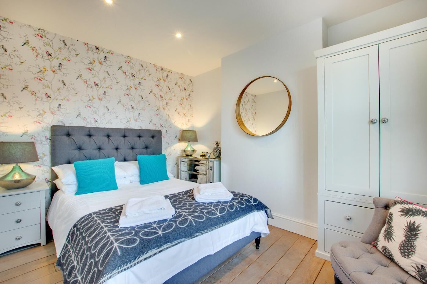 Une chambre à coucher avec une tête de lit capitonnée, un lit avec une literie blanche et bleue, un miroir rond, une armoire et une table de chevet avec une lampe.