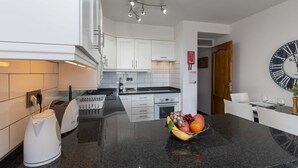 A modern kitchen with white cabinets, a black countertop, a fruit bowl, and a wall clock. - Castle Harbour 2 Bed Duplex (Los Cristianos)