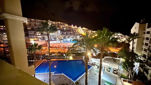 A nighttime view of a hotel with a pool, palm trees, and a busy street with cars and buildings. - Castle Harbour 2 Bed Duplex (Los Cristianos)