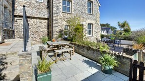 A stone building with a patio area featuring a wooden table and chairs, surrounded by potted plants and a stone wall. - 4 Bed in Padstow (oc-p00011) (Padstow)