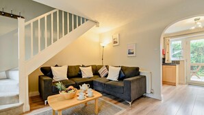 A modern living room with a grey sofa, a wooden coffee table with fruits and cups, a staircase, and a view of greenery through a glass door. - 2 Bed in Padstow (oc-p00183) (Padstow)