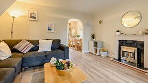 A cozy living room with a sofa, a coffee table with fruit, a fireplace, and a dining area in the background. - 2 Bed in Padstow (oc-p00183) (Padstow)