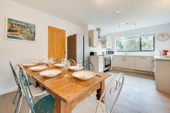 Une cuisine moderne avec des meubles blancs, une table à manger en bois pour quatre personnes et une grande fenêtre laissant entrer la lumière naturelle.