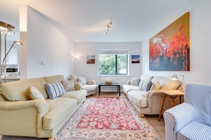 A living room with two sofas, a coffee table, a patterned rug, a painting, and a window with a view of trees. - 5 Bed in Aldeburgh (oc-pops) (Aldeburgh)