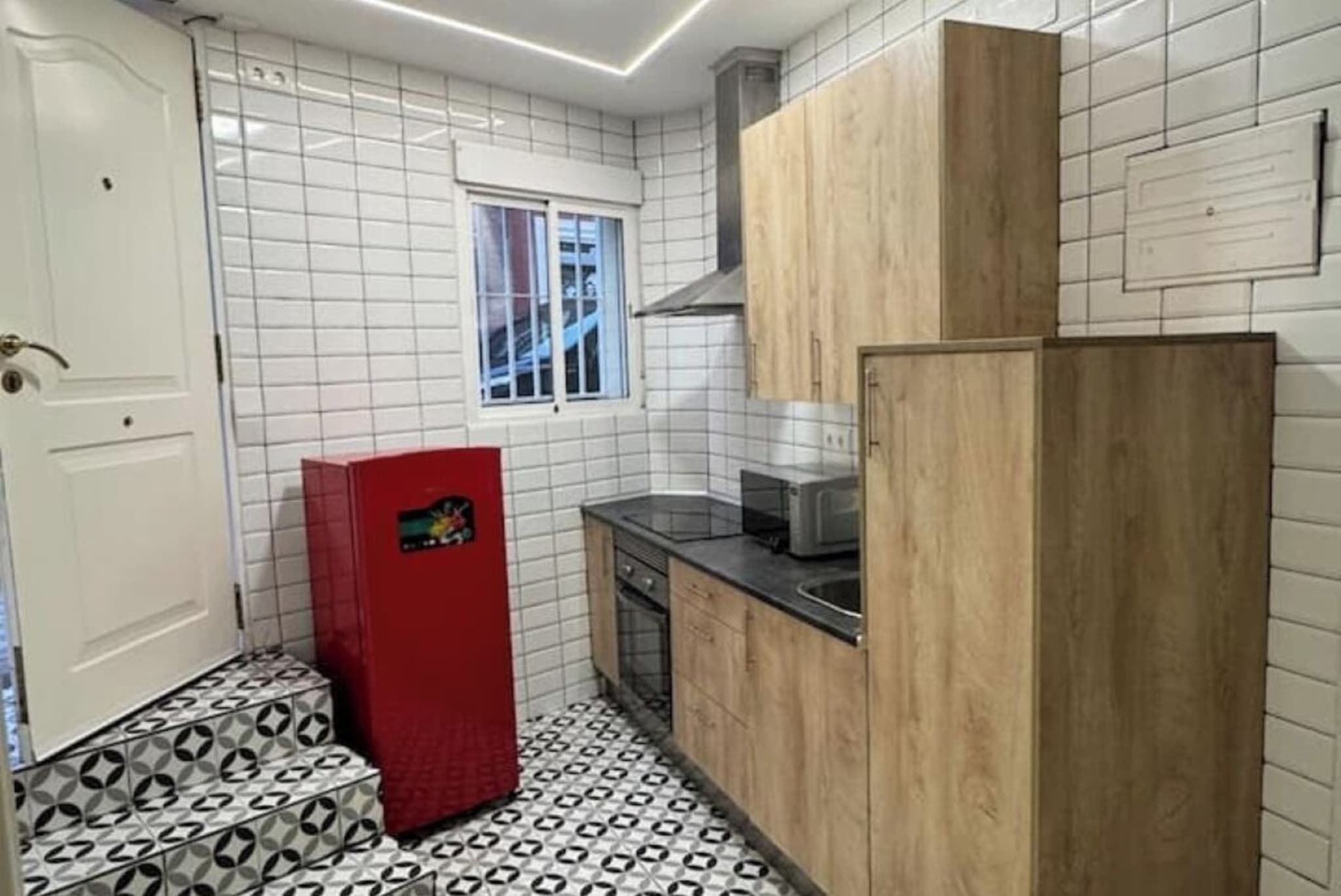 A compact kitchen with white tiled walls, a patterned floor, and wooden cabinets.