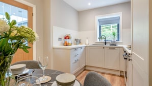 Een kleine keuken met witte kastjes, een raam en een eethoek met een tafel gedekt voor twee personen.