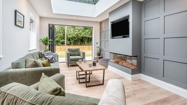 Een moderne woonkamer met een hoge plafond, een open haard, een televisie en een groot raam met uitzicht op een tuin.