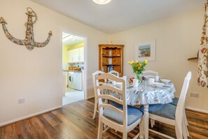 A dining area with a table set for four, wooden chairs, a chandelier, and a view into the kitchen. - 3 Bed in Padstow (oc-p00105) (Saint Merryn)