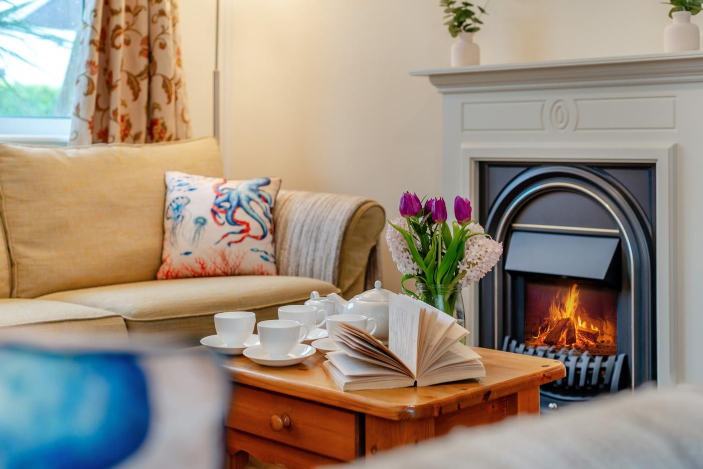 A cozy living room with a sofa, a fireplace, a coffee table with cups and books, and a vase with flowers.