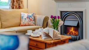 Ein gemütliches Wohnzimmer mit einer Couch, einem Kamin, einem Couchtisch mit Tassen und Büchern sowie einer Vase mit Blumen.