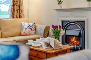 A cozy living room with a sofa, a fireplace, a coffee table with cups and books, and a vase with flowers. - 3 Bed in Padstow (oc-p00105) (Saint Merryn)