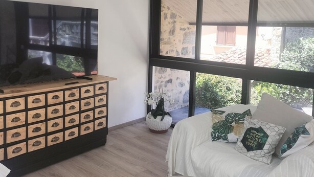 Een moderne woonkamer met een houten kast, een flatscreen televisie, een groot raam met uitzicht op een gebouw en groen, en een witte bank met decoratieve kussens.
