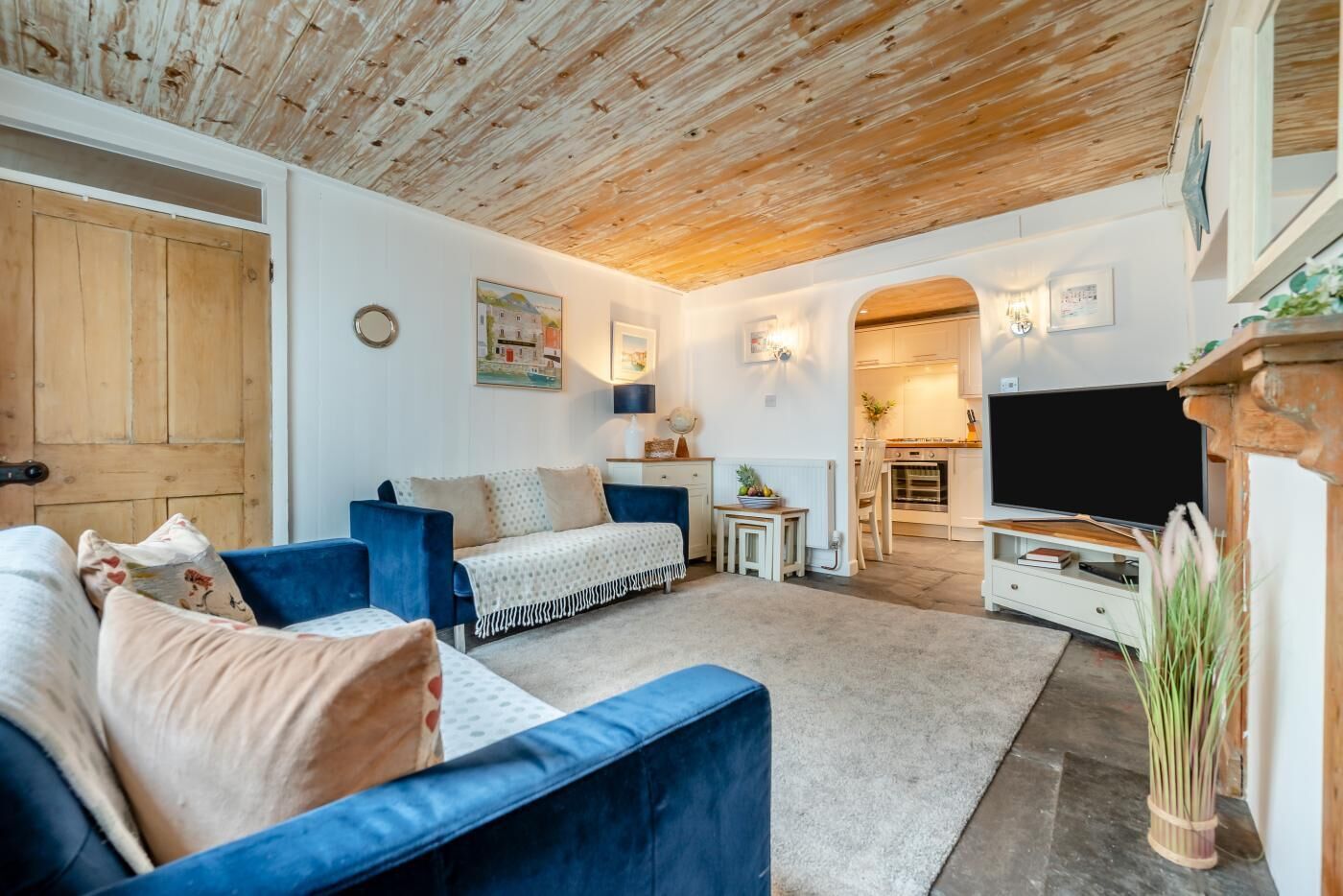 Un salon chaleureux avec un canapé bleu, une petite table basse et un téléviseur. Le plafond est en bois et on aperçoit une cuisine à travers une arche.