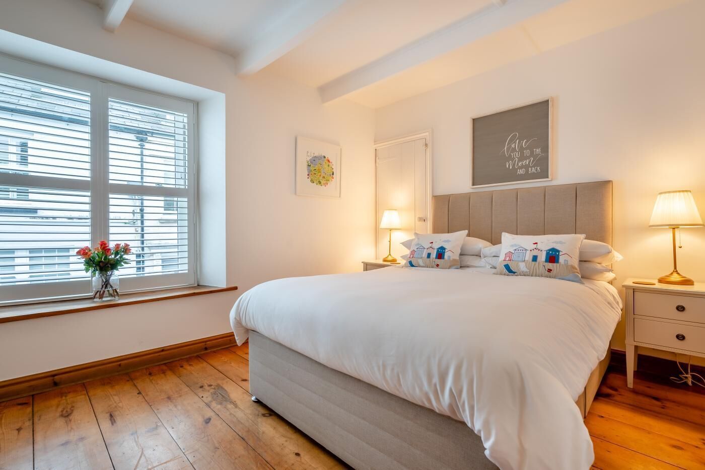 Un dormitorio con una cama grande, suelo de madera, una ventana con persianas, una mesita de noche con lámpara y un cuadro enmarcado en la pared.