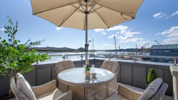Una mesa redonda con sillas de mimbre y un paraguas color beige en un balcón con vista a un puerto deportivo con barcos.