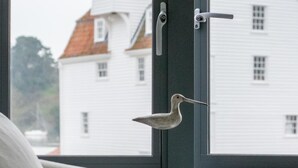 Una ventana con un pájaro posado en el pestillo, una silla en primer plano y un edificio blanco con techo rojo al fondo.