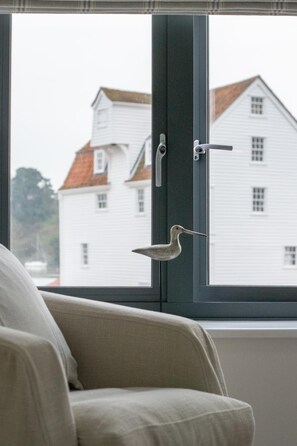 A window with a bird perched on the latch, a chair in the foreground, and a white building with a red roof in the background.