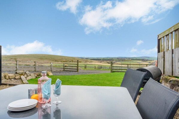A table with plates, glasses, and a bottle, set against a backdrop of a grassy field and hills. - 1 Bed in Powburn (oc-gd1919) (South Yearle)