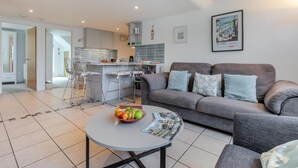 Ein modernes Wohnzimmer mit einem grauen Sofa, einem runden Couchtisch mit Obstschale und einer Küche im Hintergrund.