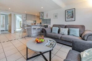 A modern living room with a grey sofa, a round coffee table with a bowl of fruit, and a kitchen in the background.