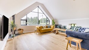 Un loft spacieux avec de hauts plafonds, de grandes fenêtres offrant une vue sur les arbres, une cheminée et un coin salon confortable avec des canapés et une table basse.