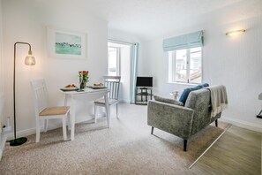 A room with a dining table, chairs, a grey sofa, a TV, and a window with curtains. - 1 Bed in St. Ives (oc-pc466a) (St Ives)