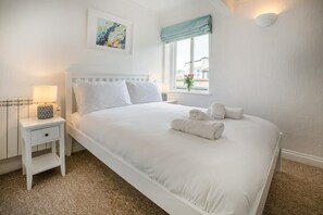 A neatly made bed with white linens and rolled towels, a bedside table with a lamp, a window with a view, and a wall-mounted light fixture. - 1 Bed in St. Ives (oc-pc466a) (St Ives)