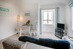 A room with a dining table set for two, a television, a window with a view of boats, and a cozy seating area with a fur throw. - 1 Bed in St. Ives (oc-pc466a) (St Ives)