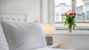 En seng med hvide sengetøj, en sengetavlelampe og et vindue med udsigt til bygninger.