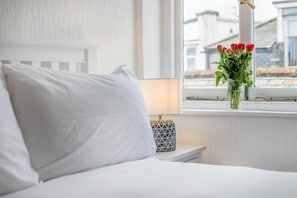 A bed with white bedding, a bedside lamp, and a window with a view of buildings. - 1 Bed in St. Ives (oc-pc466a) (St Ives)