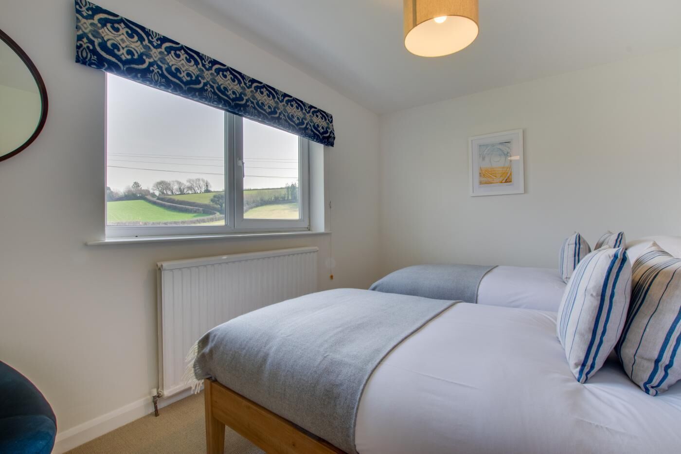 A bedroom with two beds, a window with a view of a green field, a radiator, and a ceiling light.