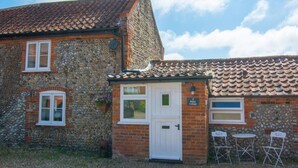 A stone cottage with a white door, two windows, and a small patio with chairs. - 1 Bed in Bodham (oc-2141) (Bodham)