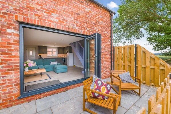 A modern brick house with a large sliding glass door leading to a living area with a sofa, a dining table, and a kitchen in the background. - 1 Bed in Marishes (oc-l32534) (Marishes)
