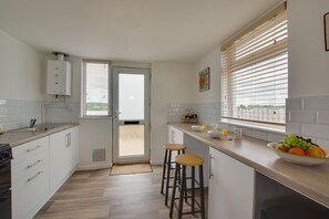 Una cocina moderna con armarios blancos, horno empotrado, un lavamanos y una ventana con persianas. Hay dos taburetes y un cuenco de fruta en la encimera.