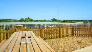 Un pontile in legno con un tavolo e due bicchieri da vino, affacciato su una superficie d'acqua tranquilla e una costa lontana.
