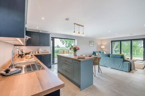 A modern kitchen with a central island, a dining area with a table and chairs, and a living room with a sofa and a view of the outdoors.