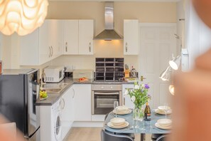 A modern kitchen with white cabinets, a built-in oven, a microwave, and a dining area with a table set for four. - 2 Bed in Padstow (oc-p00398) (Saint Merryn)