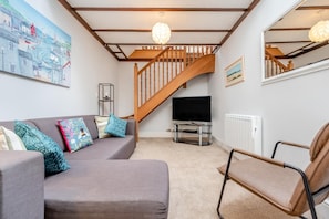 A living room with a grey sofa, a brown leather chair, a wooden staircase, a TV on a stand, and a painting on the wall. - 2 Bed in Padstow (oc-p00398) (Saint Merryn)