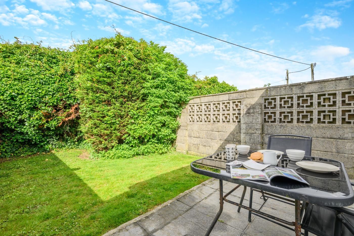 Pátio com mesa posta para refeições, cercado por um hedge verde e exuberante e uma parede de pedra com padrão.