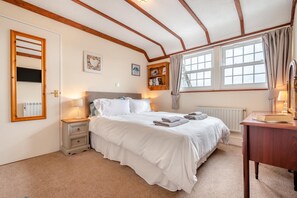 A bedroom with a bed, bedside tables, a mirror, a radiator, a window with curtains, and a bookshelf. - 2 Bed in Padstow (oc-p00398) (Saint Merryn)