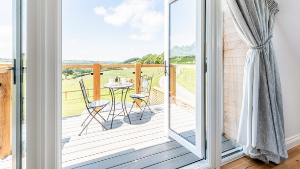 An open sliding glass door leading to a deck with a table and chairs, overlooking a grassy landscape. - 1 Bed in Umberleigh (oc-f32531) (Chittlehampton)