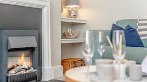 A cozy living room with a fireplace, a shelf with decorative items, and a table with glasses. - 4 Bed in Padstow (oc-p00245) (Padstow)
