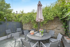 A round metal table with grey chairs, a grey umbrella, and a small table with a plate of fruit and a vase of flowers. - 4 Bed in Padstow (oc-p00245) (Padstow)