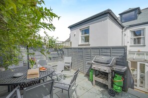 Une terrasse avec une table ronde et des chaises, un barbecue et une poubelle verte.